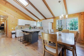 Una cocina con vigas de madera, una isla central con taburetes, armarios blancos y una mesa de comedor con sillas.