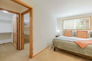 Un dormitorio con cama, mesita de noche, ventana con persianas y puerta que da a otra habitación.