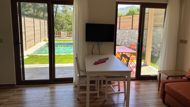 Une pièce comprenant une table blanche, un téléviseur, un canapé et une porte-fenêtre coulissante donnant sur une piscine et un espace patio.