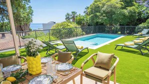 Uma área de refeições à beira da piscina, com uma mesa posta para dois, rodeada por espreguiçadeiras e vista para o mar.