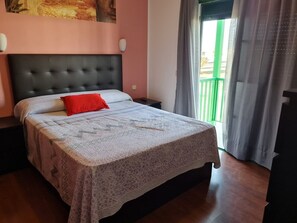 Ein Schlafzimmer mit einem Bett, einem roten Kissen, einem Kopfteil, einem Fenster mit Vorhängen und einem Bild an der Wand.