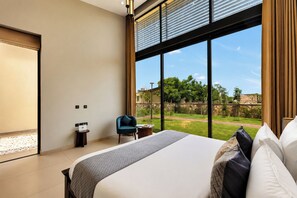 A modern hotel room with a large bed, a small table, a chair, and a view of a garden and buildings. - ELIVAAS 24 Carat 4BHK Villa with Pool (Jaipur)
