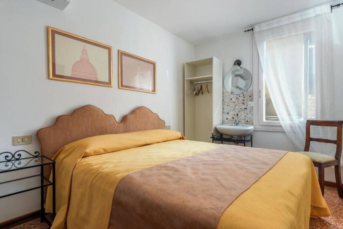 A bedroom with a bed, two framed pictures, a wardrobe, a sink, and a chair.