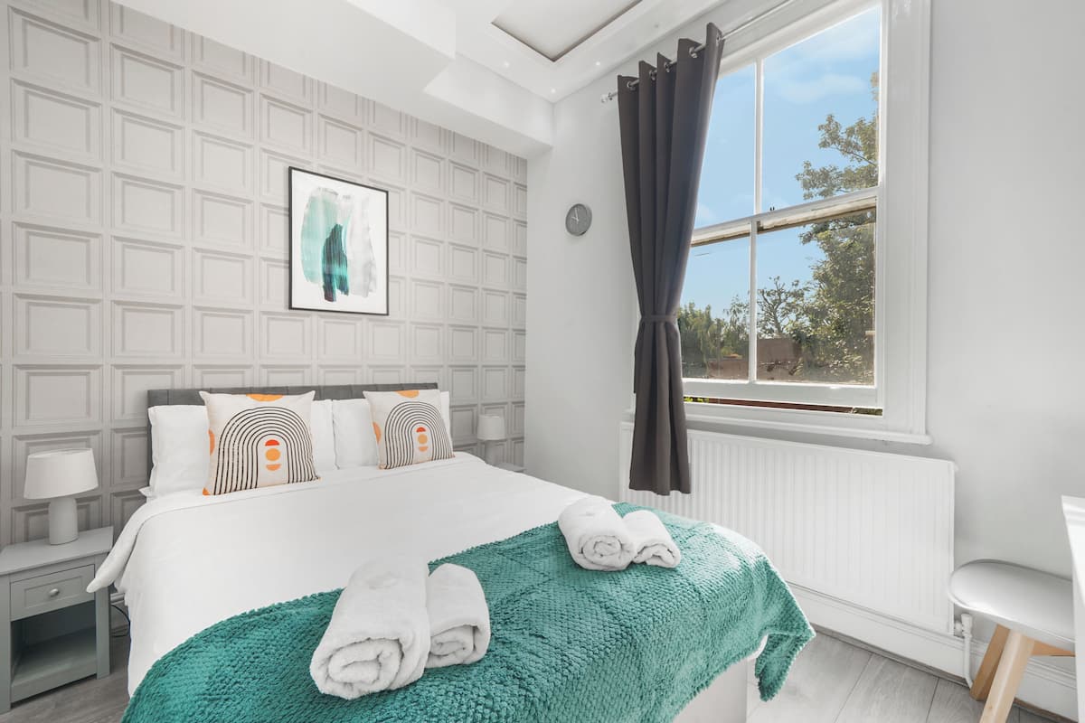 A modern hotel room with a neatly made bed, a large window with curtains, a wall clock, and a framed artwork.