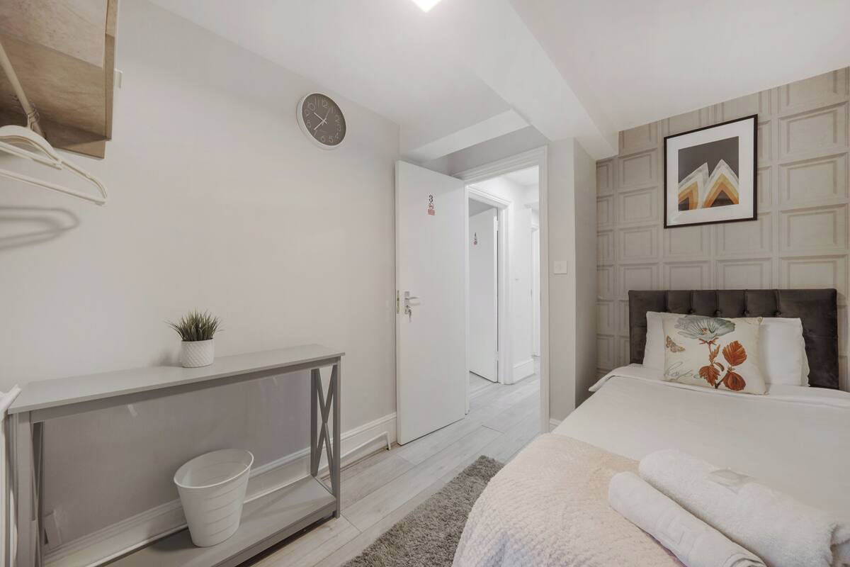 A bedroom with a bed, a bedside table, a clock, a door, and a framed picture on the wall.