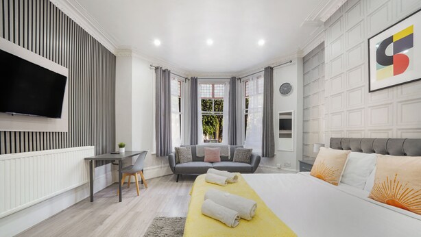 A modern hotel room with a bed, a desk, a sofa, a TV, and a large window. - The Cozy Stays Hackney House (London)