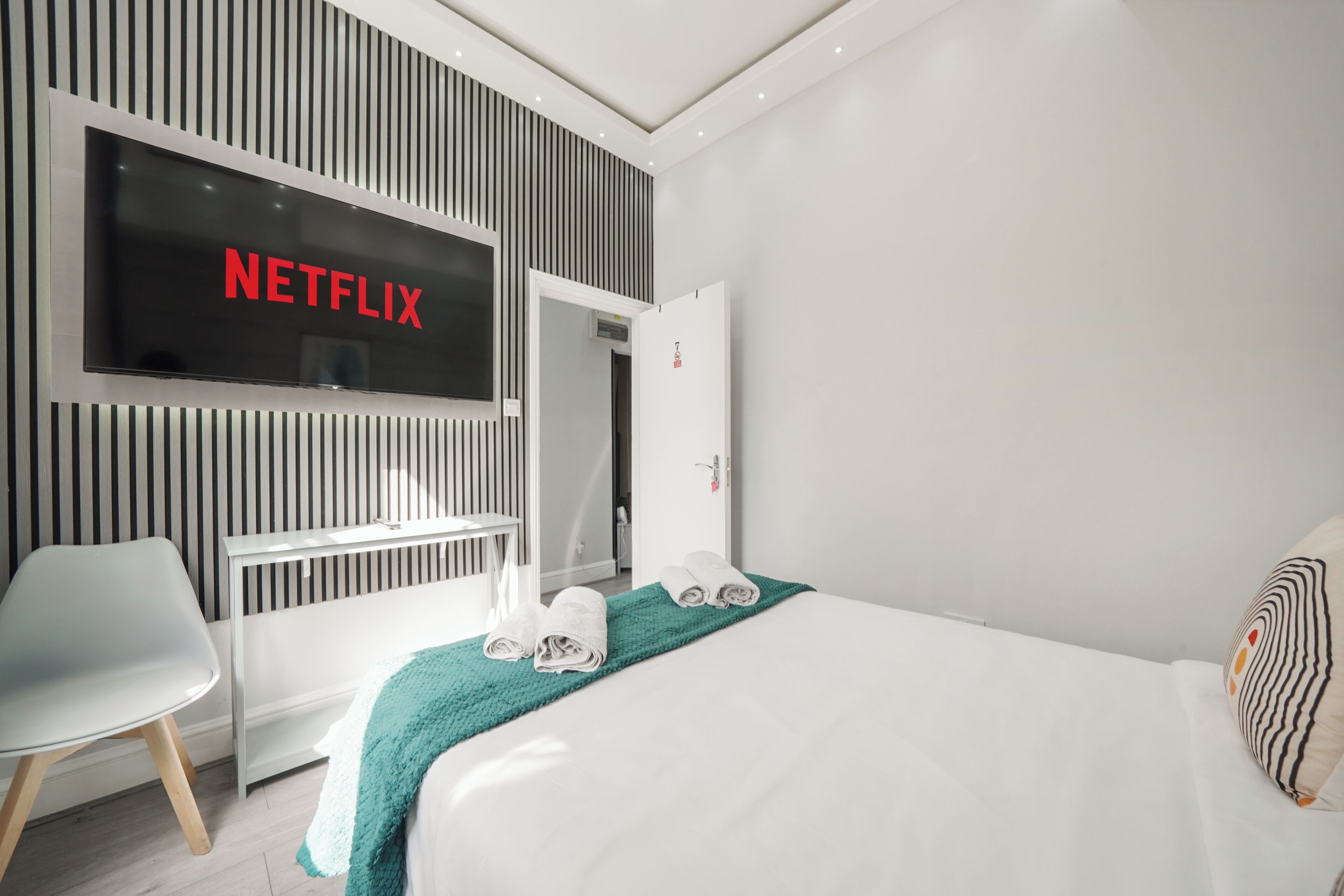 Una habitación de hotel moderna con una televisión de pantalla plana grande mostrando 'NETFLIX', una cama blanca, una manta color esmeralda y una silla blanca.