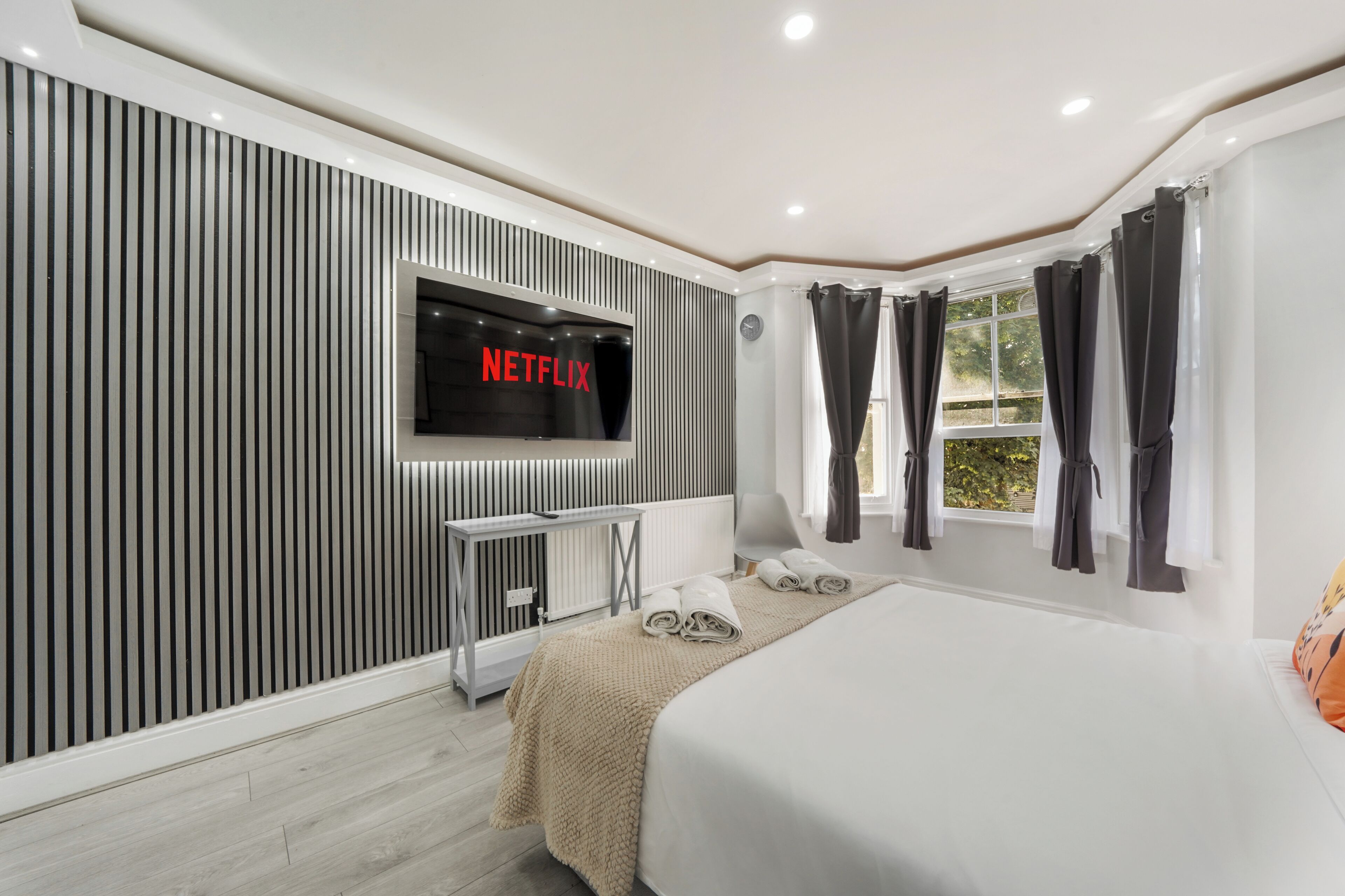 A modern hotel room with a large bed, a TV mounted on the wall, a bedside table, and a window with curtains.