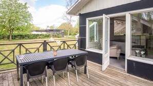 Una terraza exterior con mesa y sillas, un ventanal de vidrio que da a una habitación, y una vista de una casa al fondo.