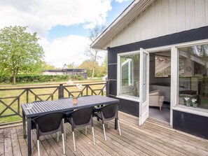 Eine Terrasse im Freien mit Tisch und Stühlen, eine Glastür, die zu einem Raum führt, und ein Haus im Hintergrund.