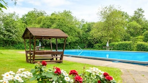 Un pabellón de madera con bancos, una piscina y un parterre de flores rojas y blancas.