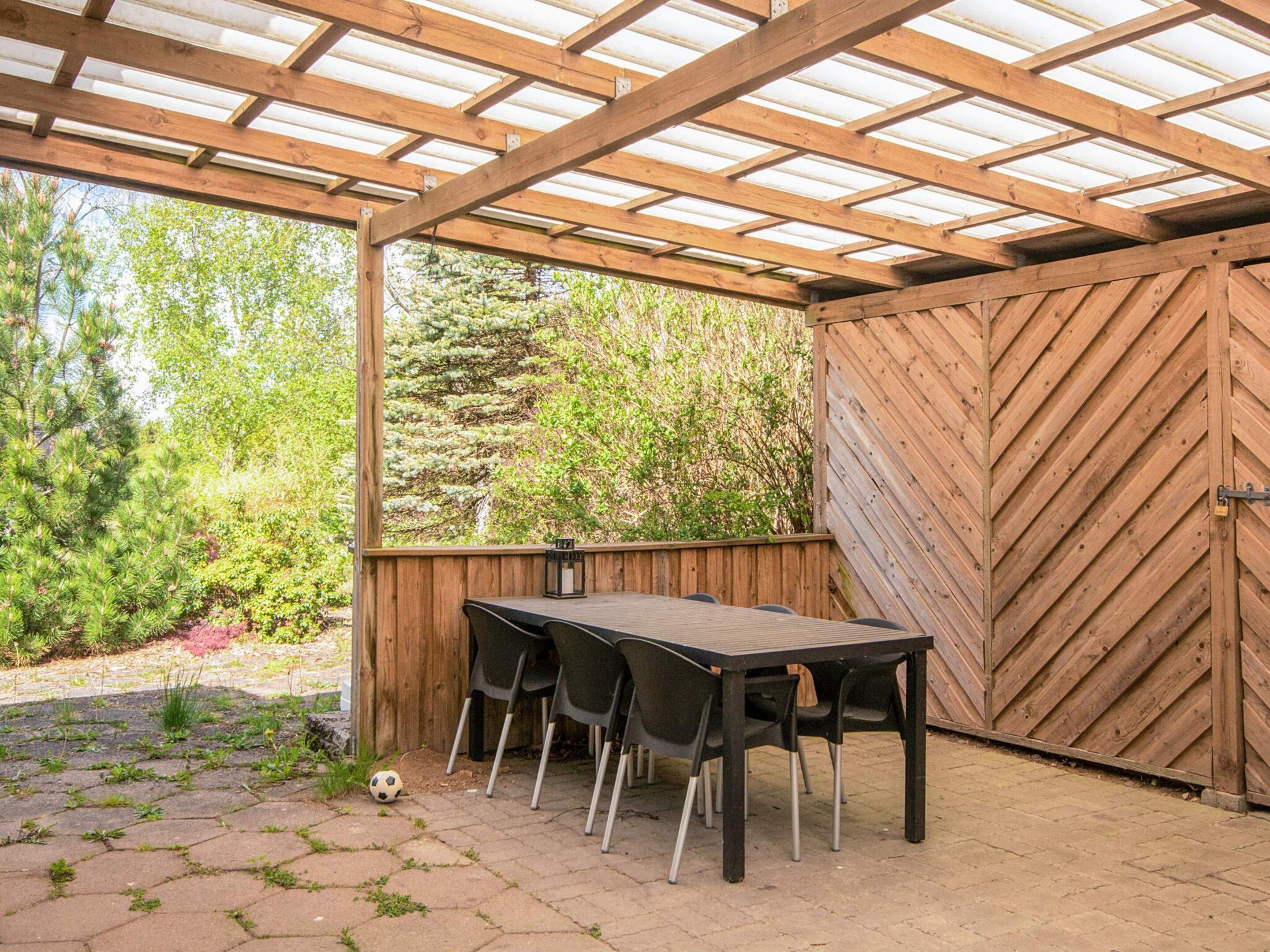 A covered patio with a wooden table and chairs, a wooden fence, and a view of greenery.