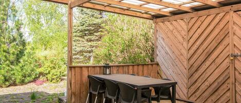 A covered patio with a wooden table and chairs, a wooden fence, and a view of greenery.