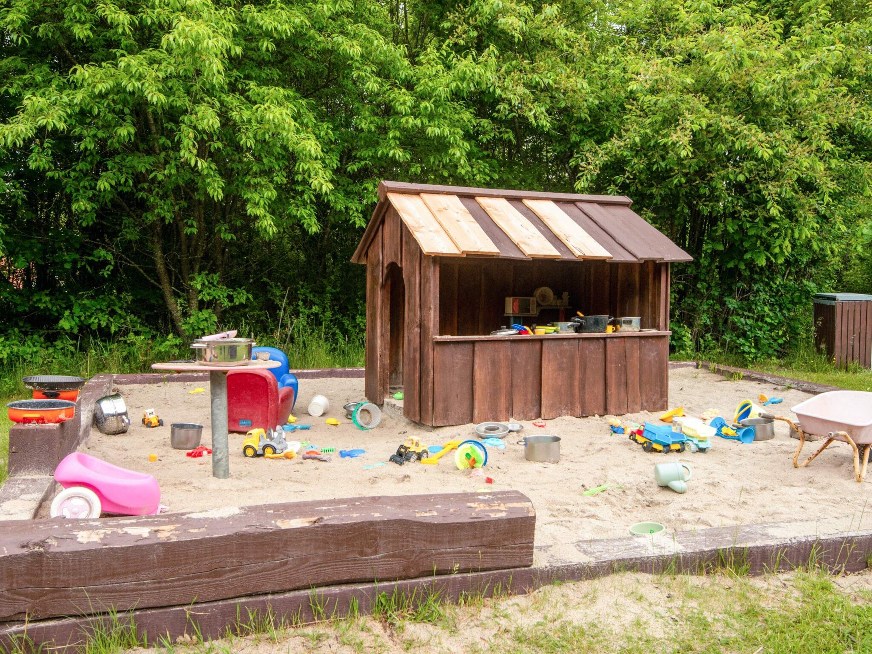 A wooden playhouse with a sloped roof, surrounded by a sandy play area with various toys and equipment.