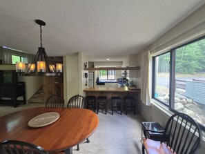 A dining area with a wooden table and chairs, a kitchen in the background, and a large window with a view of the outdoors. - Private, stand-alone, updated property, abuts woods, near Loon. C0146 by LRS (Lincoln)