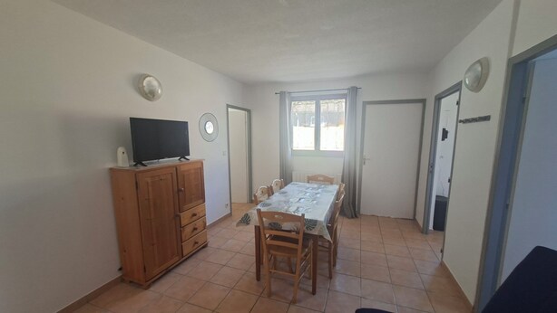 Um quarto com uma mesa de jantar, cadeiras, uma TV sobre um armário de madeira e uma porta que dá para outro cômodo.