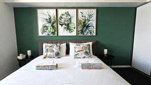 A neatly made bed with two pillows and two folded towels, a bedside table with a lamp, and three framed botanical prints on the wall. - Tropical Sunsets Studio with rooftop pool in Cairns City (Cairns City)