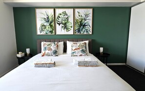 A neatly made bed with two pillows and two folded towels, a bedside table with a lamp, and three framed botanical prints on the wall. - Tropical Sunsets Studio with rooftop pool in Cairns City (Cairns City)