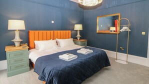 Habitación de hotel con pared azul, cabecera naranja, dos mesitas de noche con lámparas, un espejo grande y una lámpara de pie.