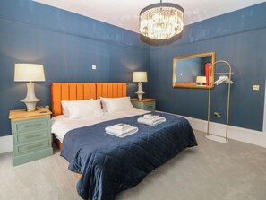 Ein Schlafzimmer mit blauer Wand, einem orangefarbenen Kopfteil, zwei Nachttischen mit Lampen, einem großen Spiegel und einer Stehlampe.