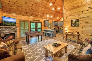 A cozy log cabin interior with a foosball table, a stone fireplace, a kitchen area, and a television. - Mountain Creek Oasis | Private Heated Indoor Pool + Hot Tub (Sevierville)