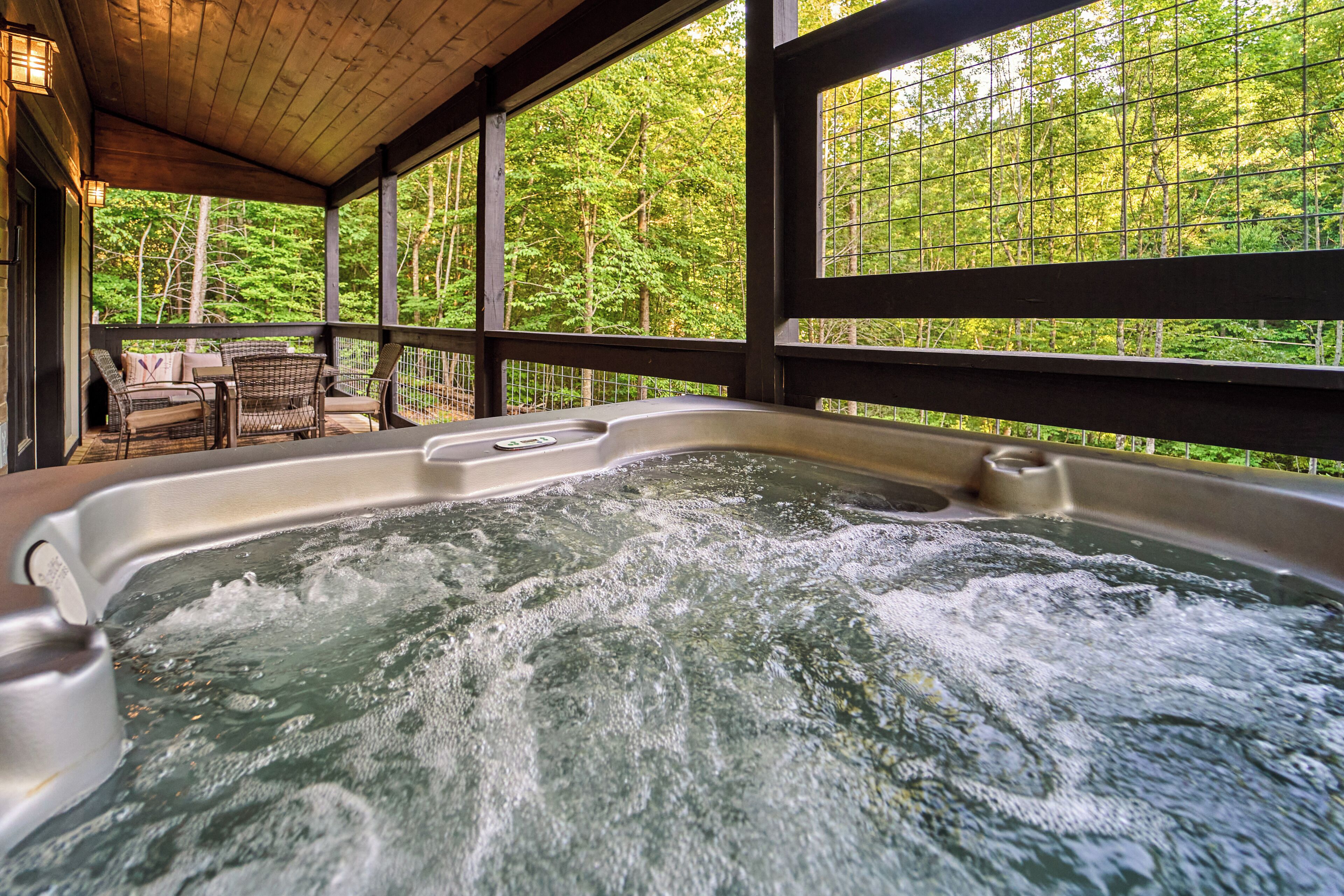 Un jacuzzi con cubierta de madera y sillas, rodeado de grandes ventanales con vista al bosque.