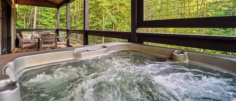 Un jacuzzi con cubierta de madera y sillas, rodeado de grandes ventanales con vista al bosque.
