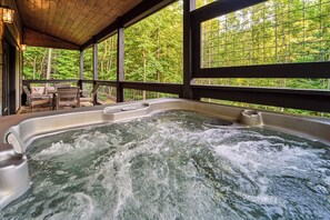 Un jacuzzi con cubierta de madera y sillas, rodeado de grandes ventanales con vista al bosque.