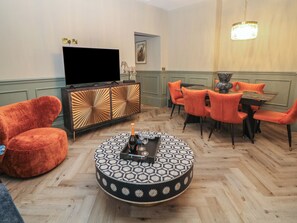 Ein Wohnzimmer mit Fernseher, einem runden Couchtisch mit Flasche und Gläsern, orangefarbenen Sesseln und einer Holzkommode.