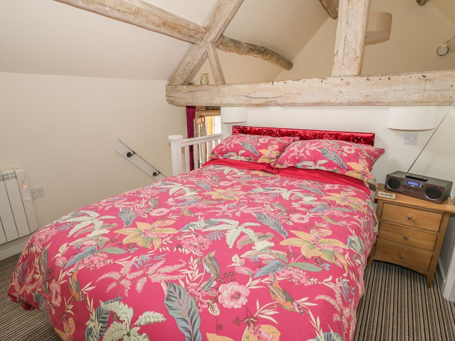 A bedroom with a floral bedspread, wooden beams, a nightstand with a radio, and a radiator.