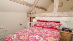 A bedroom with a floral bedspread, wooden beams, a nightstand with a radio, and a radiator. - The Bolt Hole (Stratford-upon-Avon)