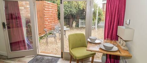 Un espacio abierto con comedor, una silla verde, una mesa preparada para dos personas y vistas del jardín a través del ventanal.