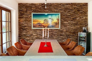 Un coin repas agrémenté d’un mur en pierre, d’un tableau représentant un paysage, d’un chandelier et de fauteuils en cuir.