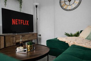 Een woonkamer met een televisie waarop 'NETFLIX' staat, een houten kast, een groene bank, een ronde salontafel en een wandklok.