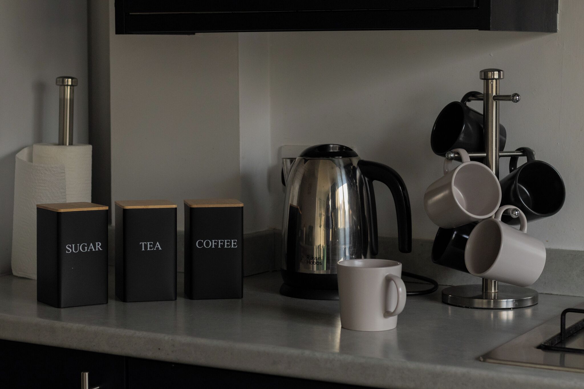 Een keukenblad met daarop gemarkeerde potten voor suiker, thee en koffie, een waterkoker en een kopjeshouder met kopjes.