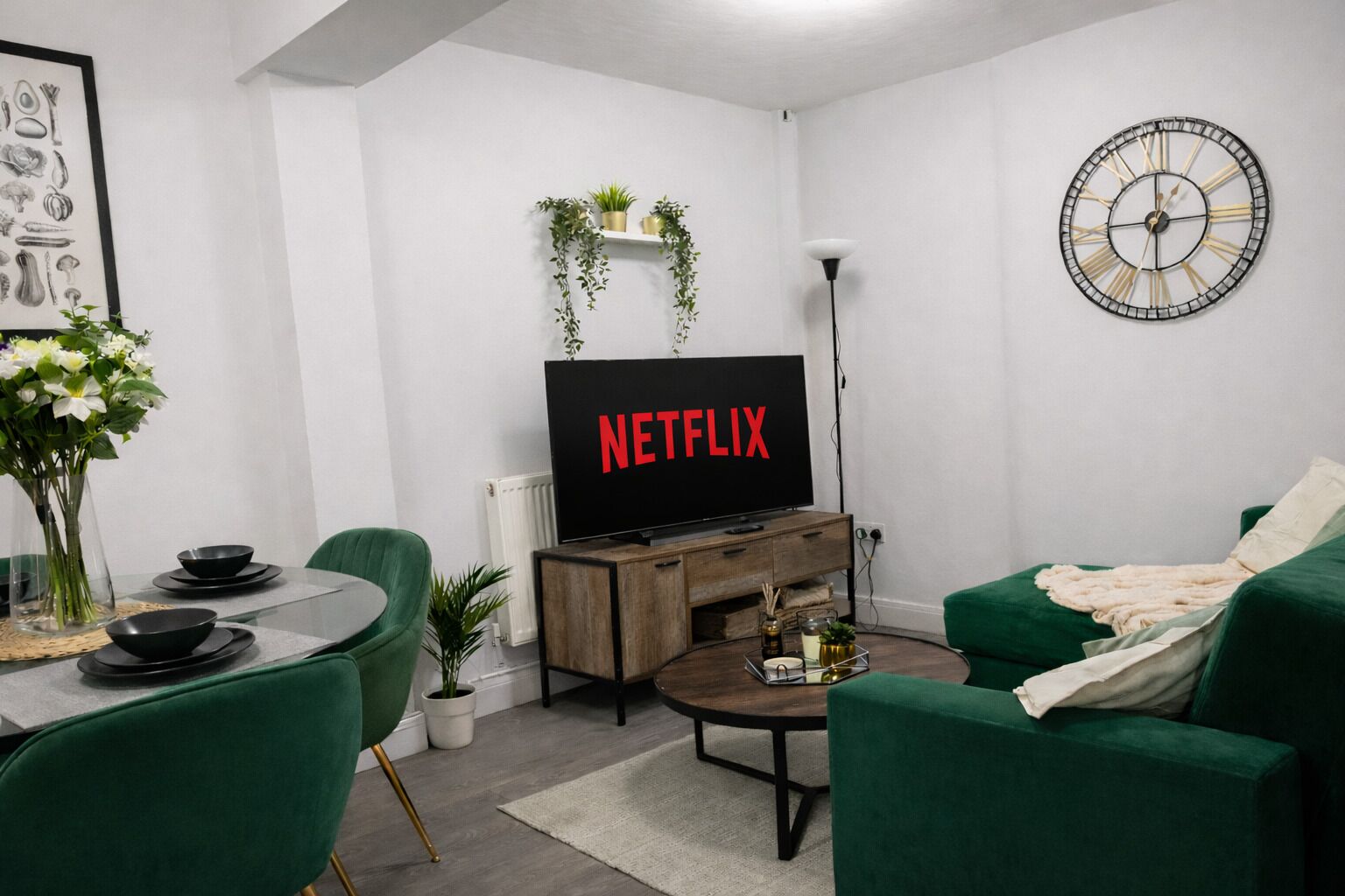 A modern living room with a TV displaying 'NETFLIX,' a green sofa, a coffee table, a dining area with a glass table, chairs, and a vase of flowers.