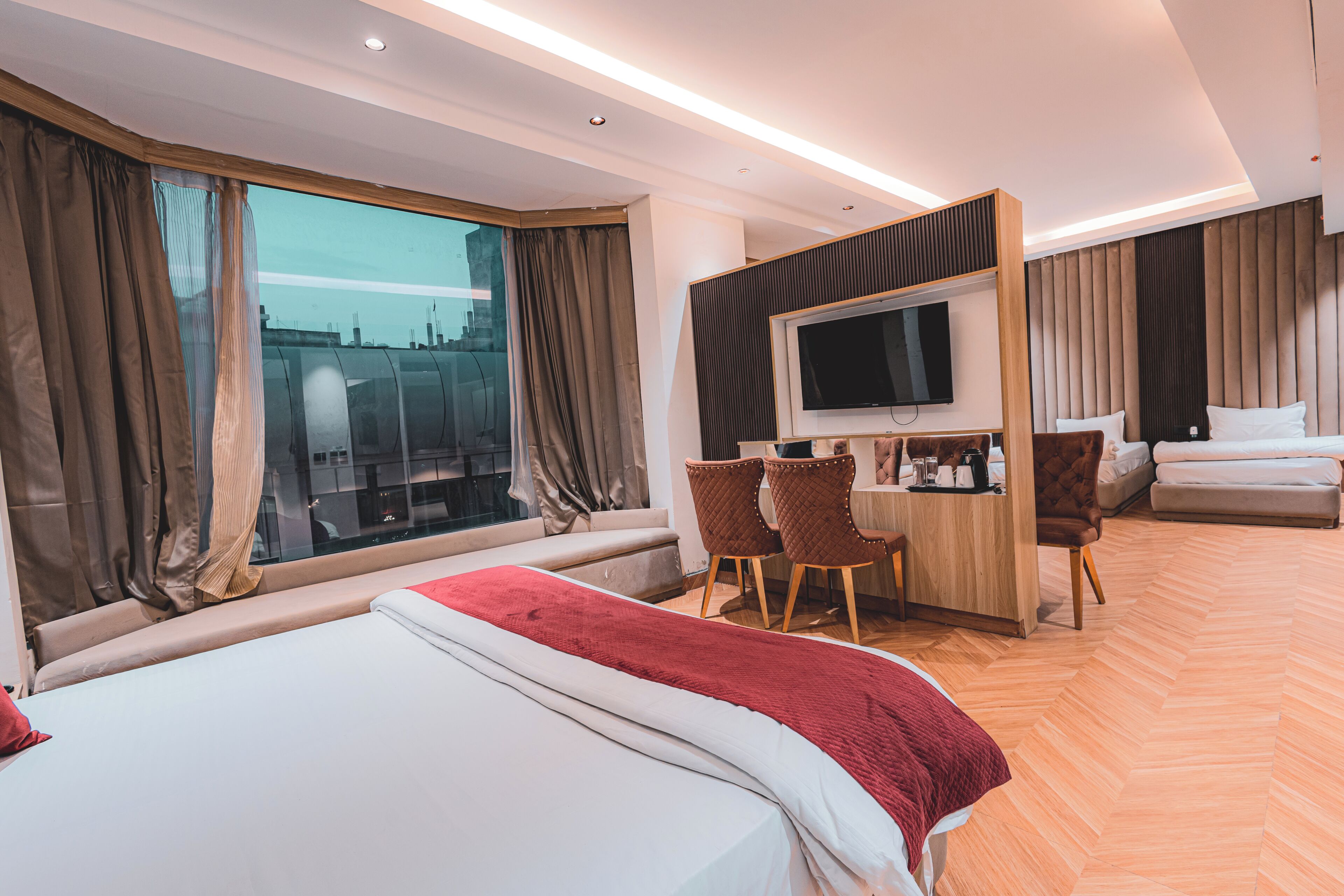 A hotel room with a bed, a desk with a TV, a seating area, and a view of the city.