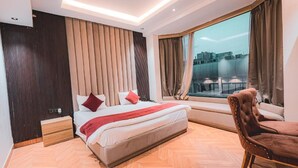 Quarto de hotel moderno com uma cama grande, janela com vista para a cidade, piso de madeira e uma poltrona de couro marrom.