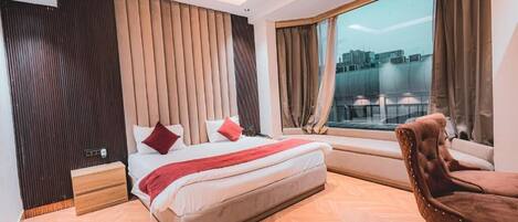 Una habitación de hotel moderna con una cama grande, un ventanal con vista a la ciudad, piso de madera y un sillón de cuero marrón.
