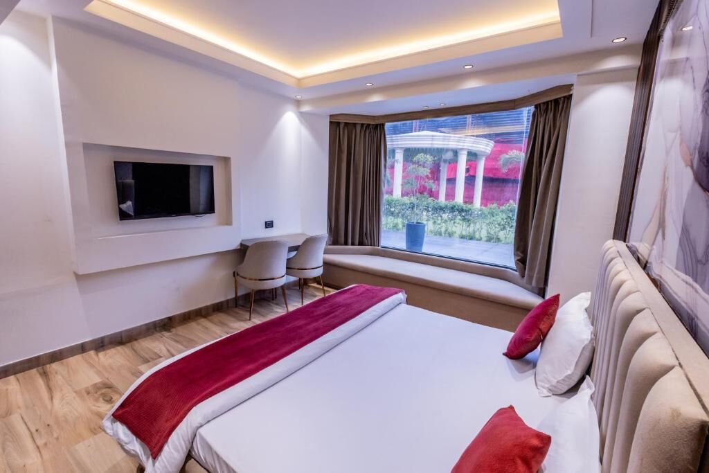 A modern hotel room with a large bed, a TV mounted on the wall, a small seating area, and a view of a garden through a window.