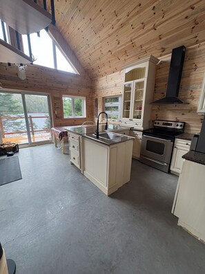 A wooden cabin kitchen with a central island, a stove, and a large window overlooking a forested area.