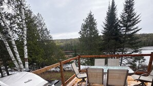 Um deck de madeira com uma churrasqueira, duas cadeiras e vista para um lago e floresta.