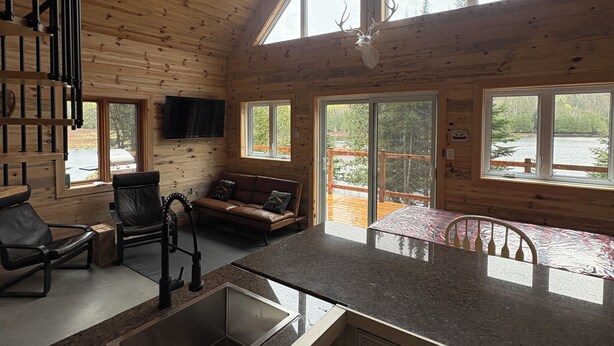Interior espaçoso de uma cabana, com cozinha, sala de jantar e uma janela grande com vista para um deck.