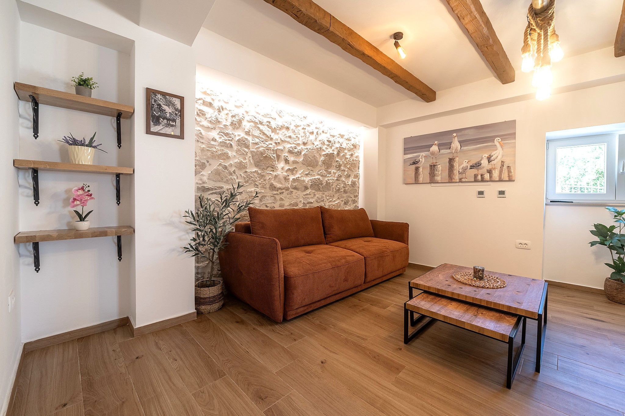 Uma sala de estar acolhedora com parede de pedra, vigas de madeira, um sofá marrom, mesa de centro de madeira e prateleiras com plantas.