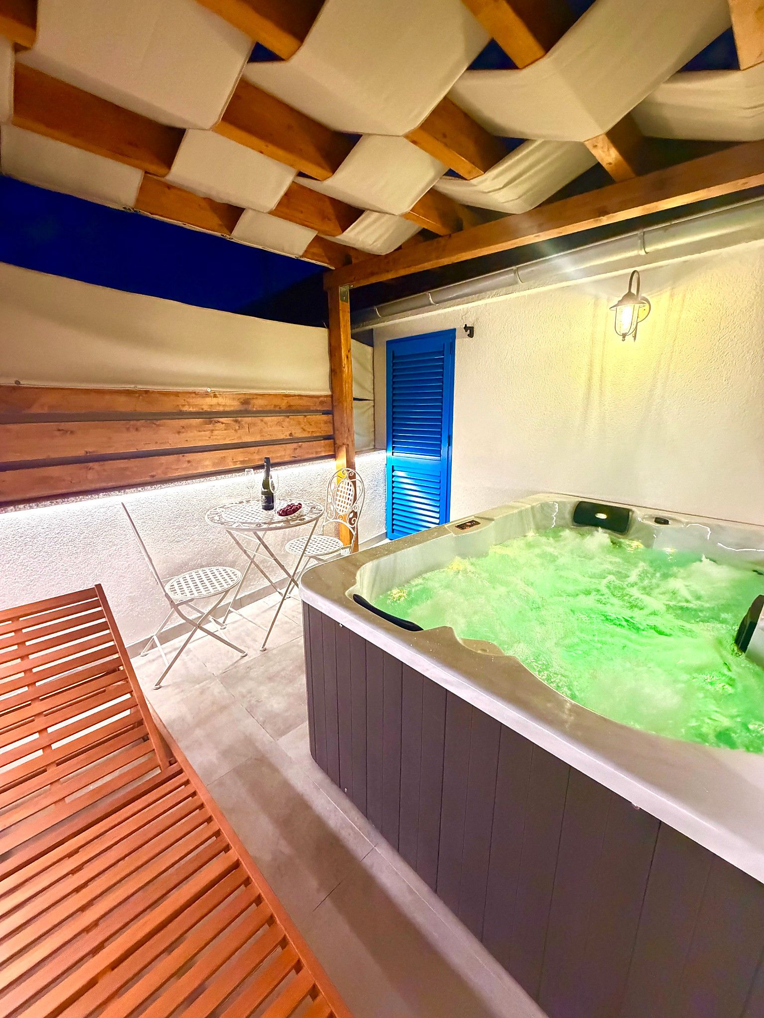 A hot tub area with a wooden bench, a table with chairs, and a blue door.