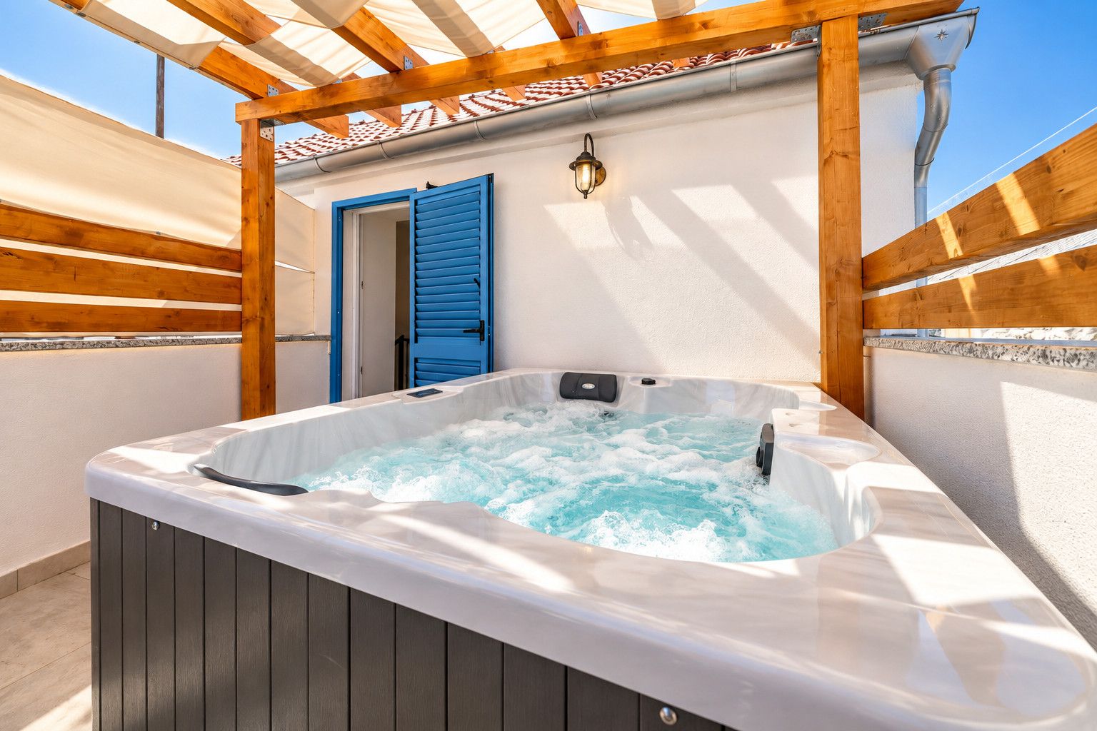 A hot tub under a wooden pergola with a blue door and a wall-mounted light fixture.