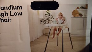Um bebê sentado em uma cadeirinha de alimentação com uma planta ao lado.