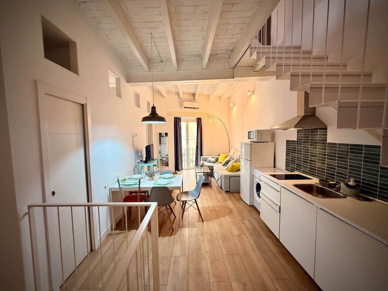 Een moderne keuken met witte kastjes, een zithoek met tafel en stoelen, en een trap die naar een verdieping leidt.