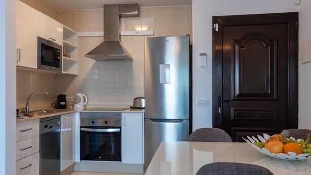 Eine kleine Küche mit Mikrowelle, Backofen und Kühlschrank. Ein Esstisch mit Stühlen und einer Obstschale steht bereit.
