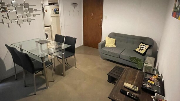 Kompakti olohuone, jossa on lasinen ruokapöytä, sohva ja pieni puinen pöytä, jonka päällä on kaukosäädin.
